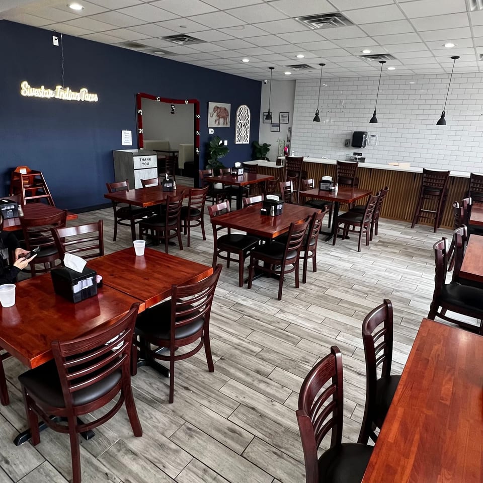 This is a wide-angle interior shot of a dining room featuring dark wood tables and chairs, light-colored tile flooring, and a blue accent wall with a neon sign that reads 'Sweetie Indian Tacos'.