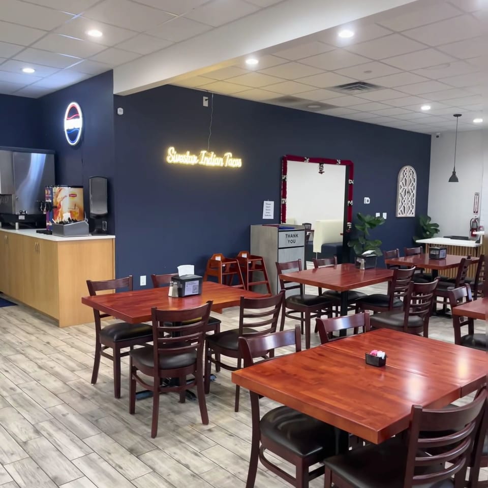 This is a real photograph of the dining area of a restaurant named 'Siestars Indian Tacos', featuring wooden tables, dark chairs, a beverage station, and a neon sign on a navy blue wall.
